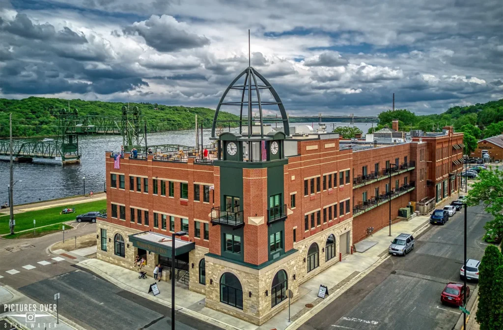 An aerial view of a brick building with a clock tower and dome next to a river with a bridge under a cloudy sky. Water Street Inn is one of the best Hotels in Stillwater