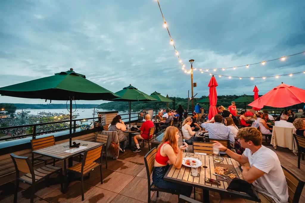 People sit at outdoor tables with umbrellas on a rooftop deck overlooking a river at dusk, with string lights overhead.