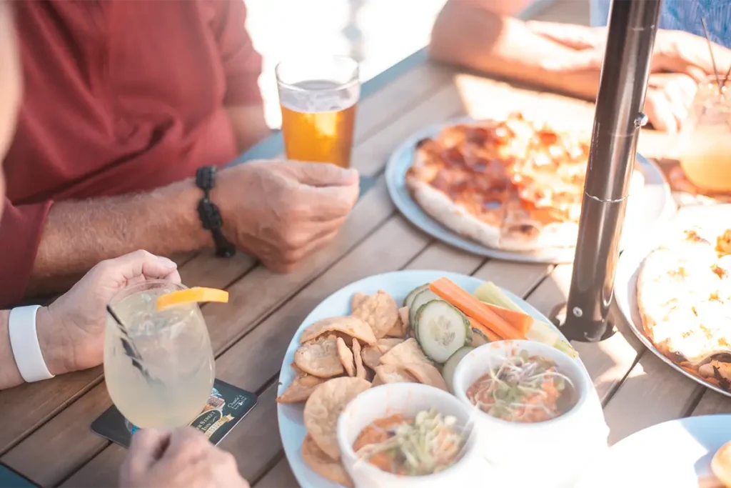 People share drinks, pizza, and a plate of pita chips with vegetables and dip on a wooden table.