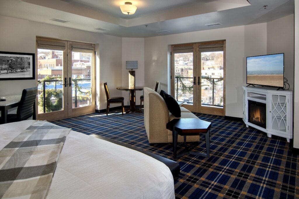 A hotel room with plaid carpet, a white bed, a small sofa, an electric fireplace with a TV, and glass balcony doors.