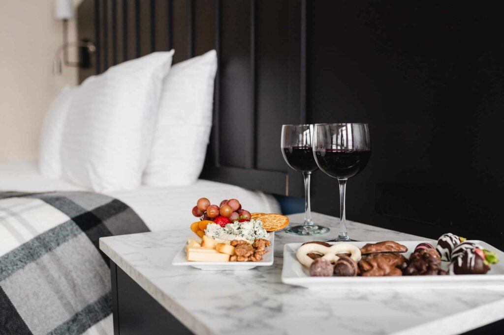 Two glasses of red wine, cheese, and chocolate treats on a marble bedside table next to a hotel bed.