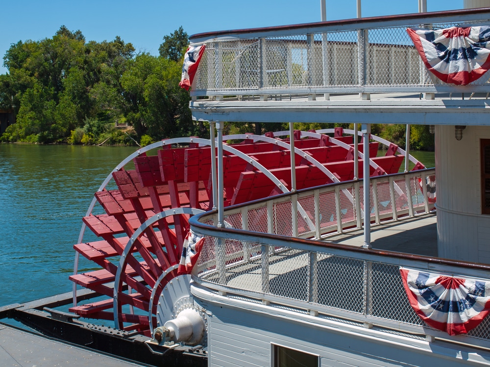 Close-up of a paddlewheel boat's rear paddle wheel and two-story deck with red, white, and blue bunting on a river. Stillwater River Boats