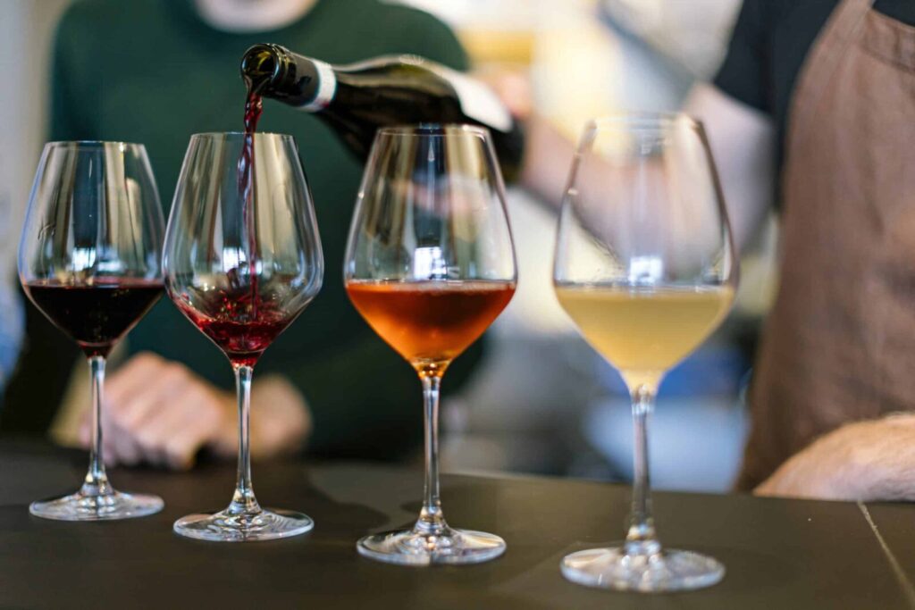 Red wine is being poured from a bottle into a line of glasses containing red, rosé, and white wine.