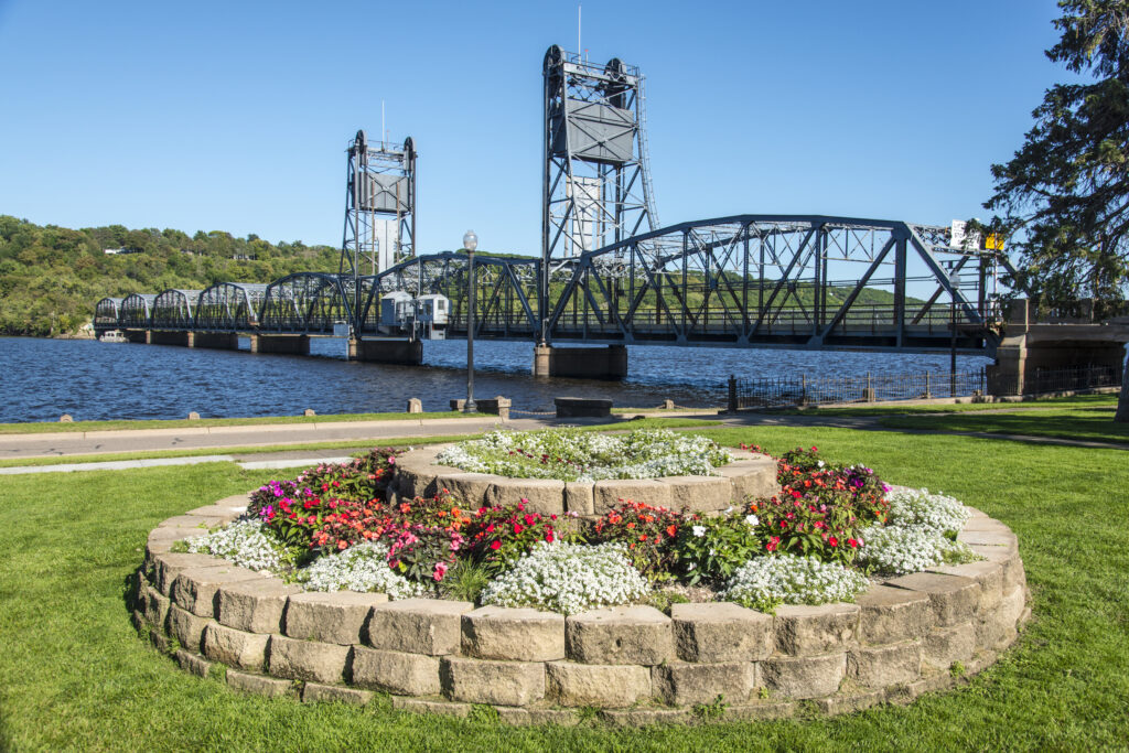 A stone flower bed sits in a park with a steel vertical lift bridge over a river in the background.