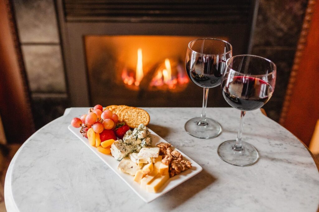 A plate of cheese, grapes, and crackers next to two glasses of red wine on a table before a glowing fireplace.